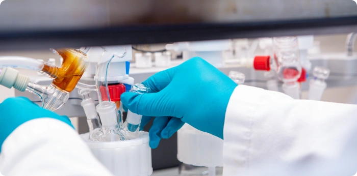 Scientist wearing blue nitrile gloves adjusting glassware apparatus during pharmaceutical chemical synthesis and formulation in a laboratory setting