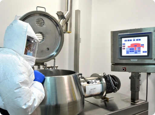 Operator in a white cleanroom hazmat suit monitoring a stainless steel high shear granulator mixer and industrial control panel during API formulation