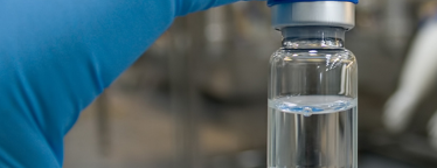 A gloved hand in a cleanroom holding up a small glass vaccine vial