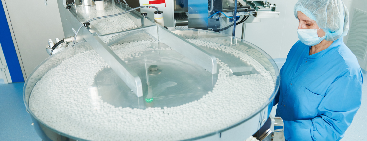 A pharmaceutical factory operator wearing a hairnet and gown monitoring an automated machine sorting hundreds of white pills on a rotating tray
