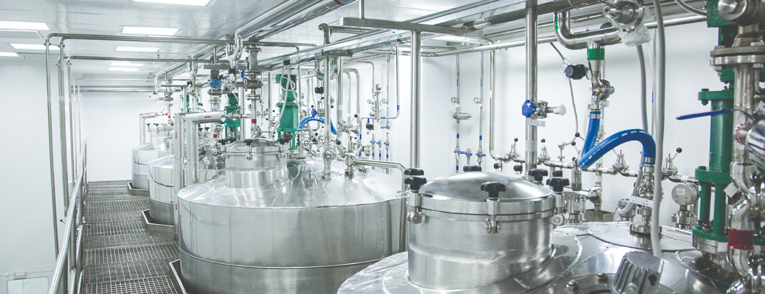 Rows of large, industrial stainless steel tanks, bioreactors, and complex piping systems inside a clean pharmaceutical manufacturing facility